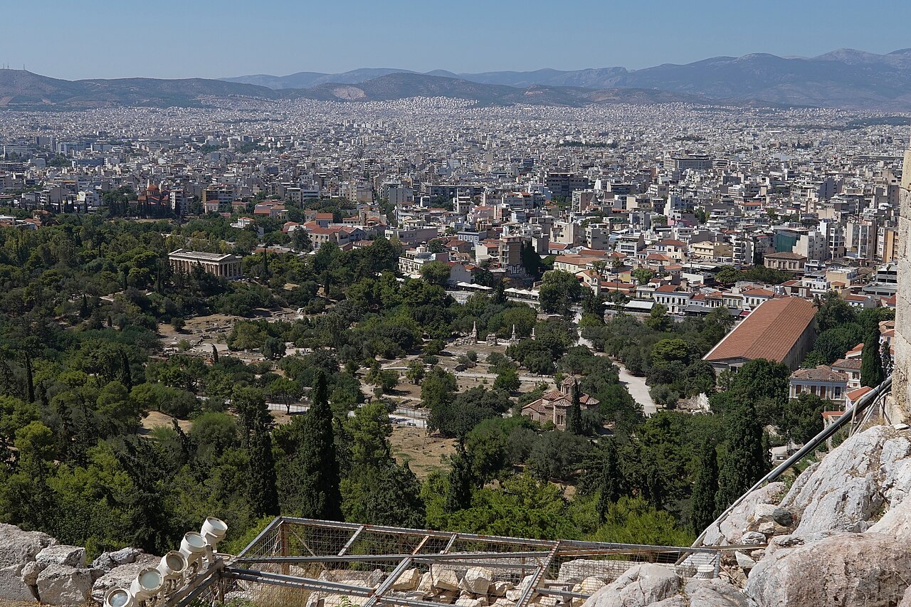 A wide view of the Ancient Agora of Athens with open ground, trees, ruins, the Stoa of Attalos, and the modern city around it.